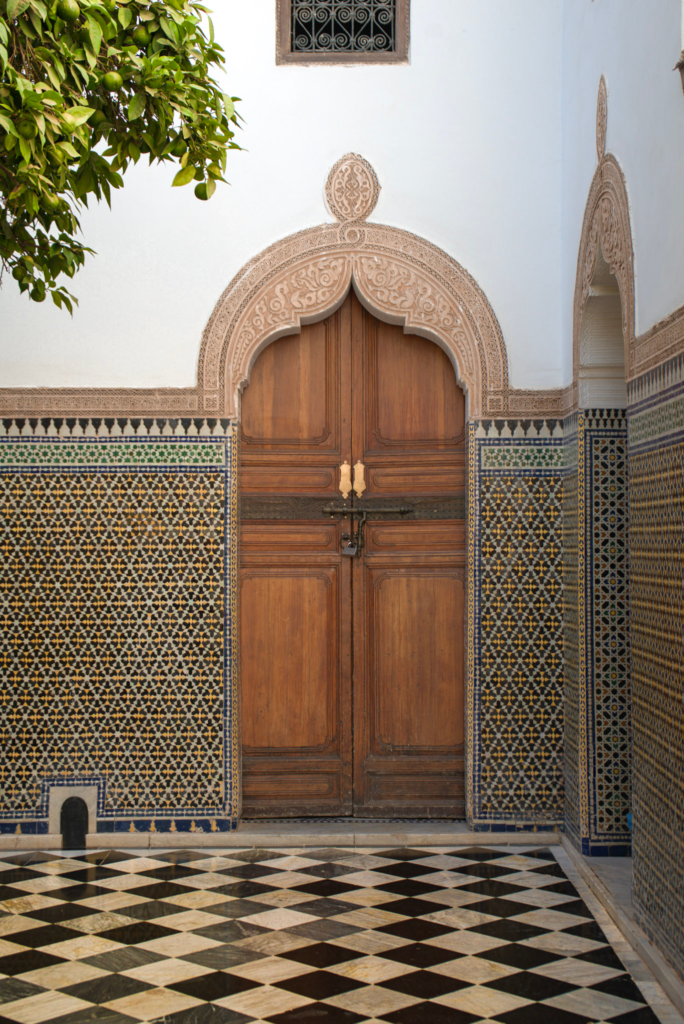Ornate cedar wood doorway at Dar El Bacha in Marrakesh framed by carved stucco arches and colorful zellij tilework, with a black and white checkered floor leading into the quiet museum courtyard. A leafy orange tree branch hangs overhead, capturing the calm architectural beauty that makes this a worthwhile stop when exploring things to do in Marrakesh.
