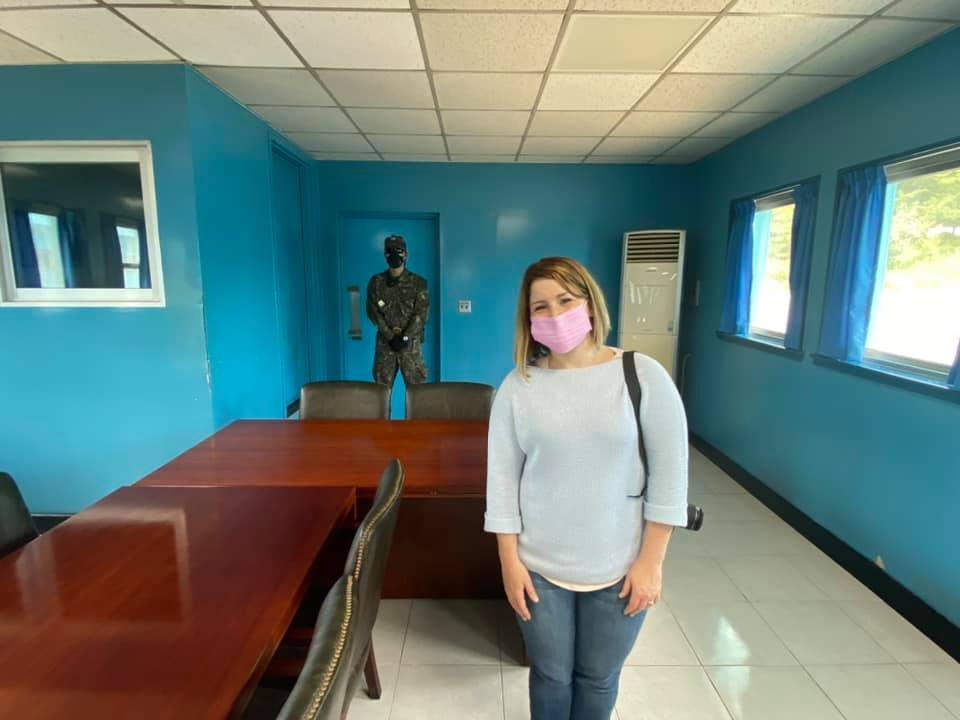 Woman wearing a pink face mask standing inside the blue Joint Security Area conference room with a soldier positioned near the door behind her.