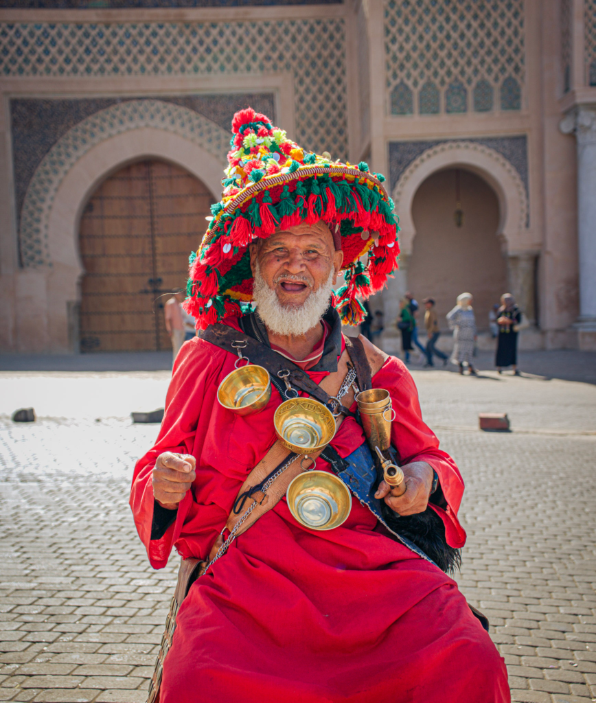 a man in a red robe holding cups and with a decorative hat in Meknes a traditional water man of Morocco