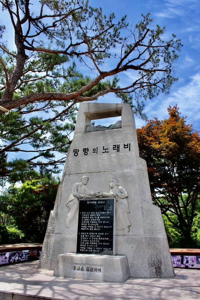 Tall stone monument engraved with Korean text and relief carvings beneath a pine tree honoring hopes for reunification near the DMZ.