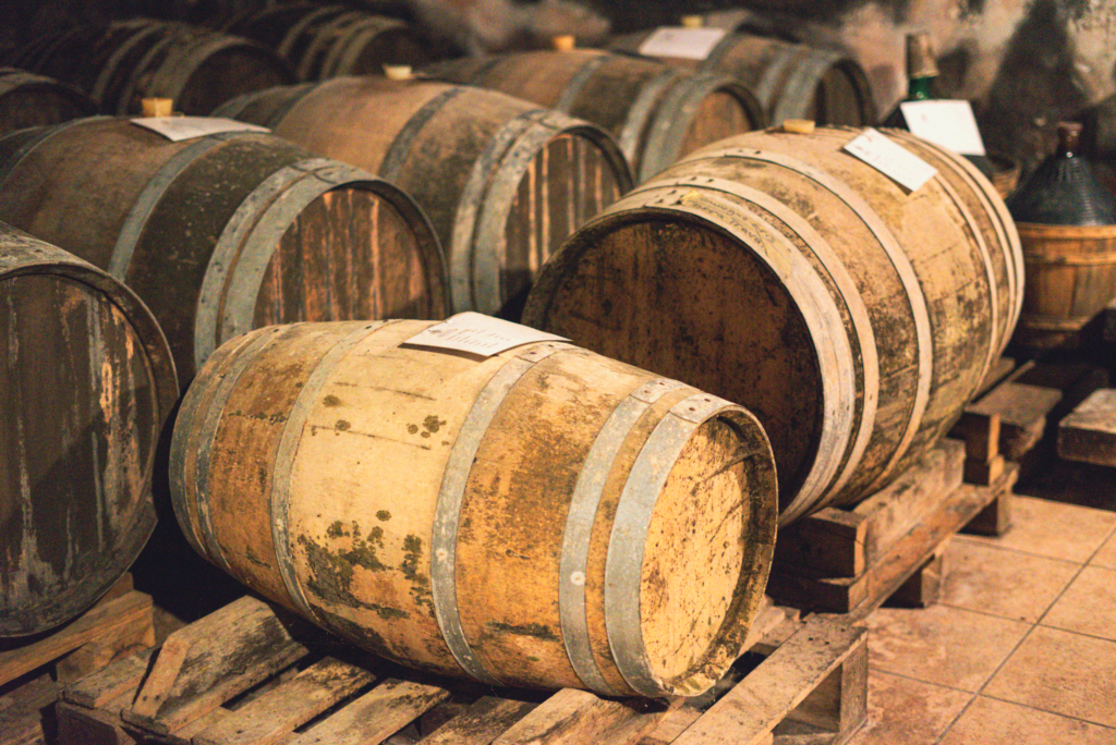 Rows of aging oak barrels rest on wooden pallets inside the cellar at Menti Wines Gambellara with warm light highlighting the textured wood and stone walls. The barrels reflect traditional aging methods at this biodynamic winery Italy known for native yeast wine and sweet wine Gambellara.