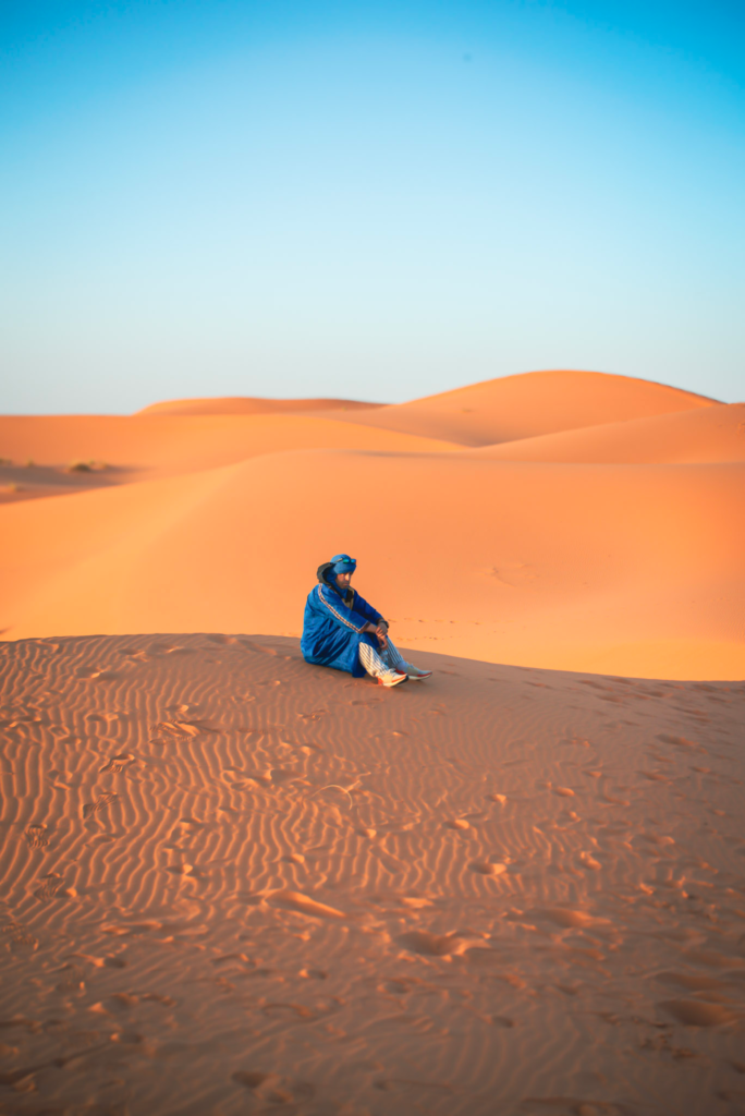 Alt text. A person dressed in a blue robe sits quietly on a rippled sand dune surrounded by vast golden desert dunes under a clear blue sky. The image captures a peaceful moment in the Moroccan desert highlighting the scale and stillness of the landscape.