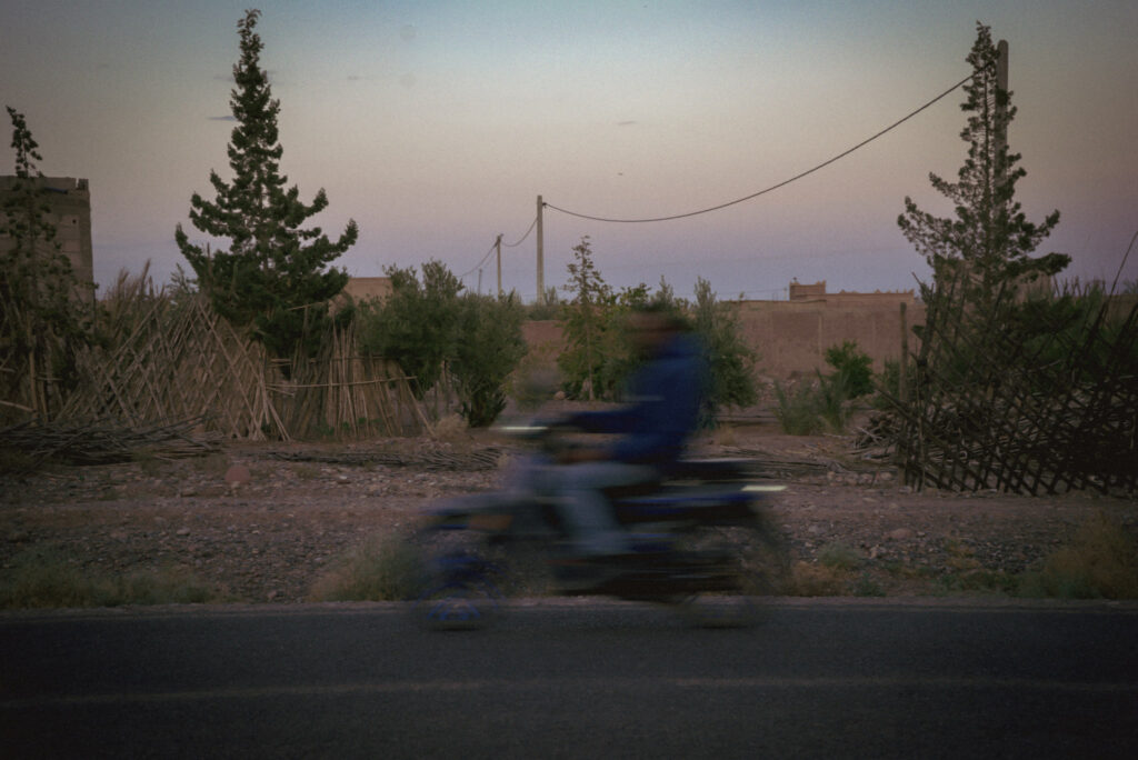 A blurred motorbike passes along a road at dusk in Skoura with palm trees wooden fencing and low buildings in the background. The motion blur captures everyday life during evening travel through southern Morocco.