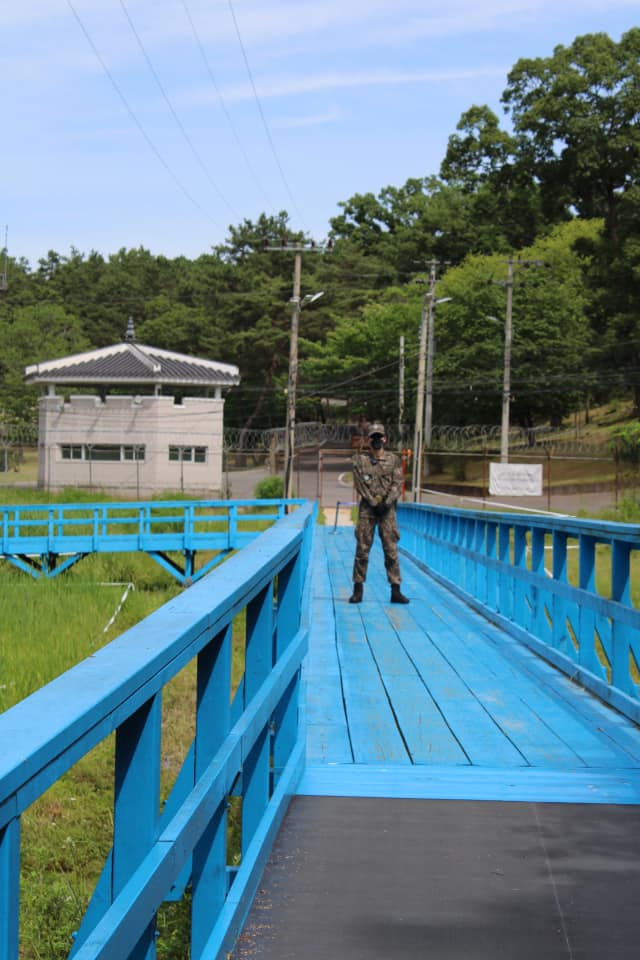 Soldier standing guard on the bright blue bridge at the Joint Security Area with barbed wire fencing and guard posts in the background.