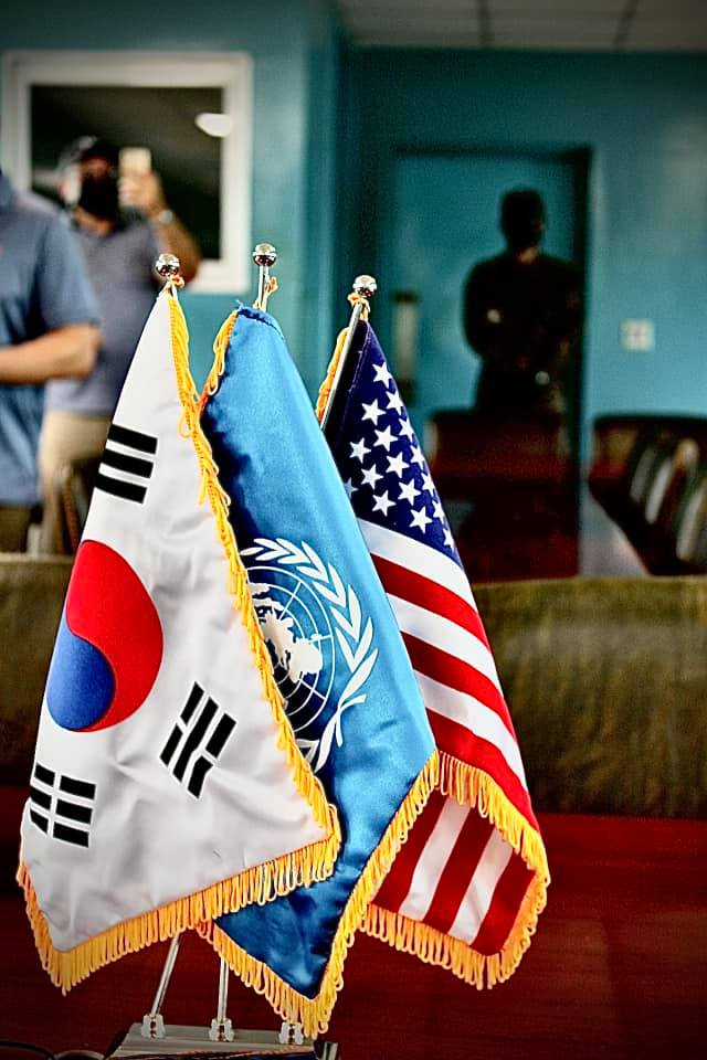 Tabletop display of South Korea United Nations and United States flags inside the Joint Security Area conference room with masked visitors reflected in the background.