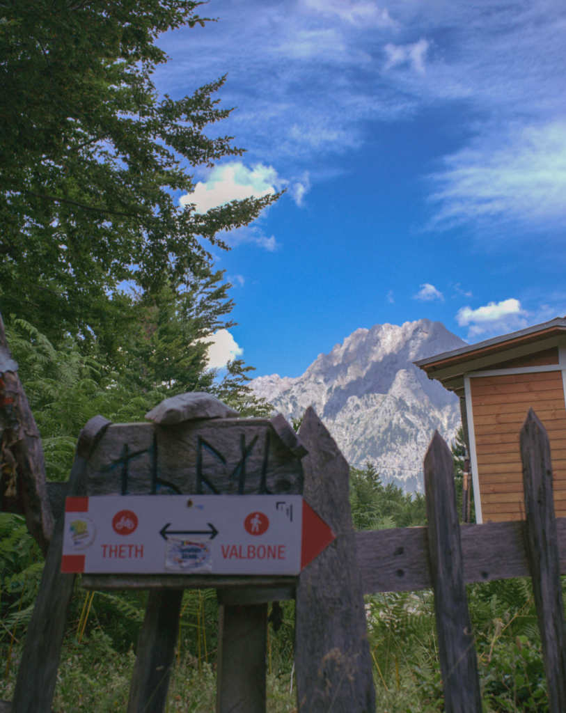 Wooden signpost reading Theth and Valbone with directional arrows and hiking symbols. The sign is mounted on a rustic fence with rugged mountains visible in the distance. It marks the start of the popular Theth to Valbone hiking trail in Albania.