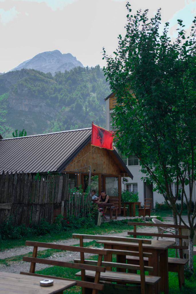 Outdoor seating area beside a wooden guesthouse with an Albanian flag waving above the roof. Mountains rise in the background while two people sit at a table near a rustic fence. The image captures village life in Theth before hiking toward Valbone Valley.