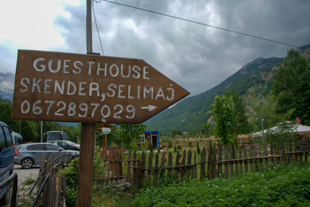 Wooden arrow sign that reads Guesthouse Skender Selimaj with the phone number 0672897029 pointing right. Parked cars and a fenced yard sit beneath cloudy mountain skies. This sign directs hikers to accommodation in Theth Valley.