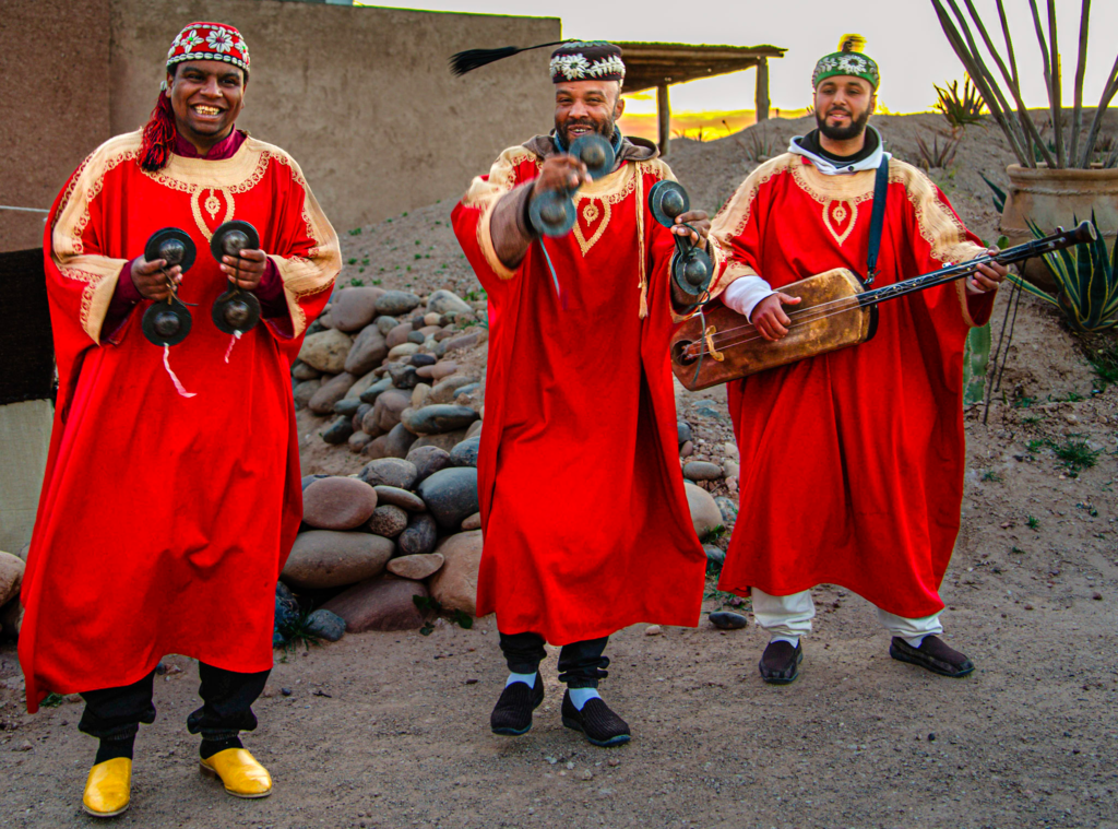 Three musicians in bright red traditional robes play castanets and a string instrument while smiling and welcoming guests at dinner at the Agafay Desert. The scene captures the lively atmosphere and cultural entertainment that greets visitors arriving for an evening meal in the desert.