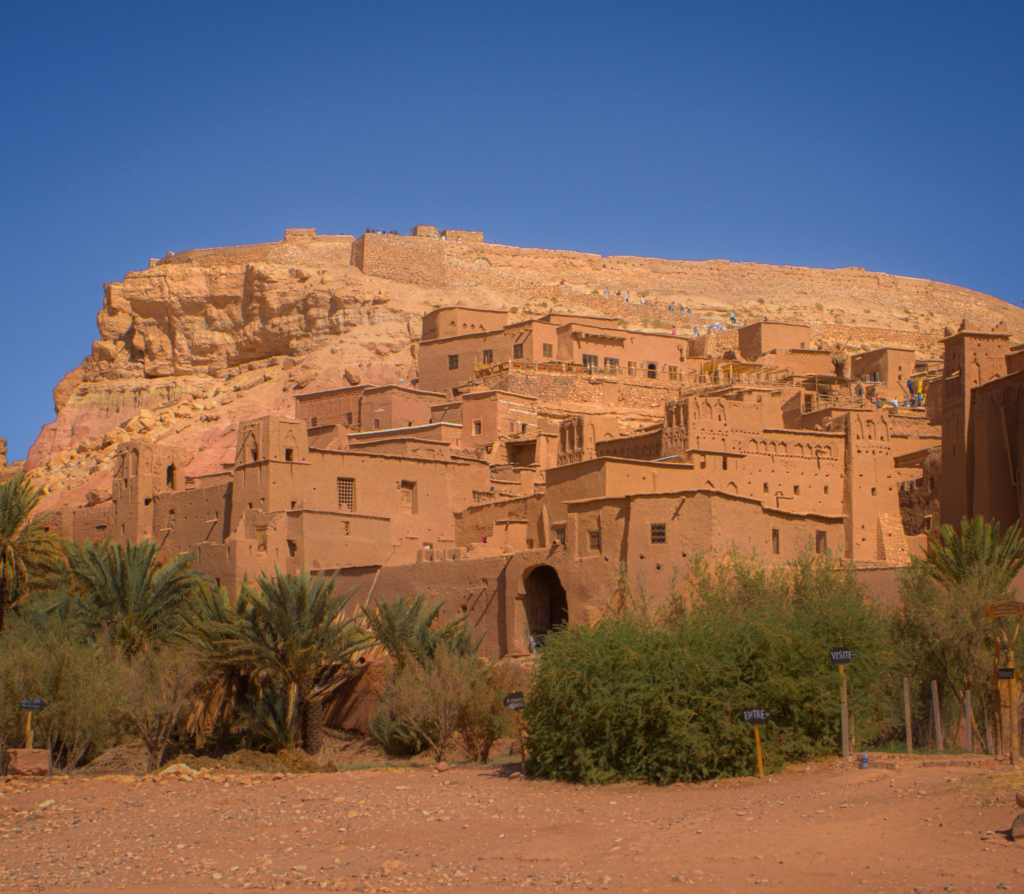 raditional earthen clay buildings of a fortified village sit at the base of a rocky hill under a clear blue sky, with palm trees and desert shrubs in the foreground. Small signs near the entrance read "VISITE" and "ENTRE," suggesting a tourist site often visited as a day trip from Marrakesh.