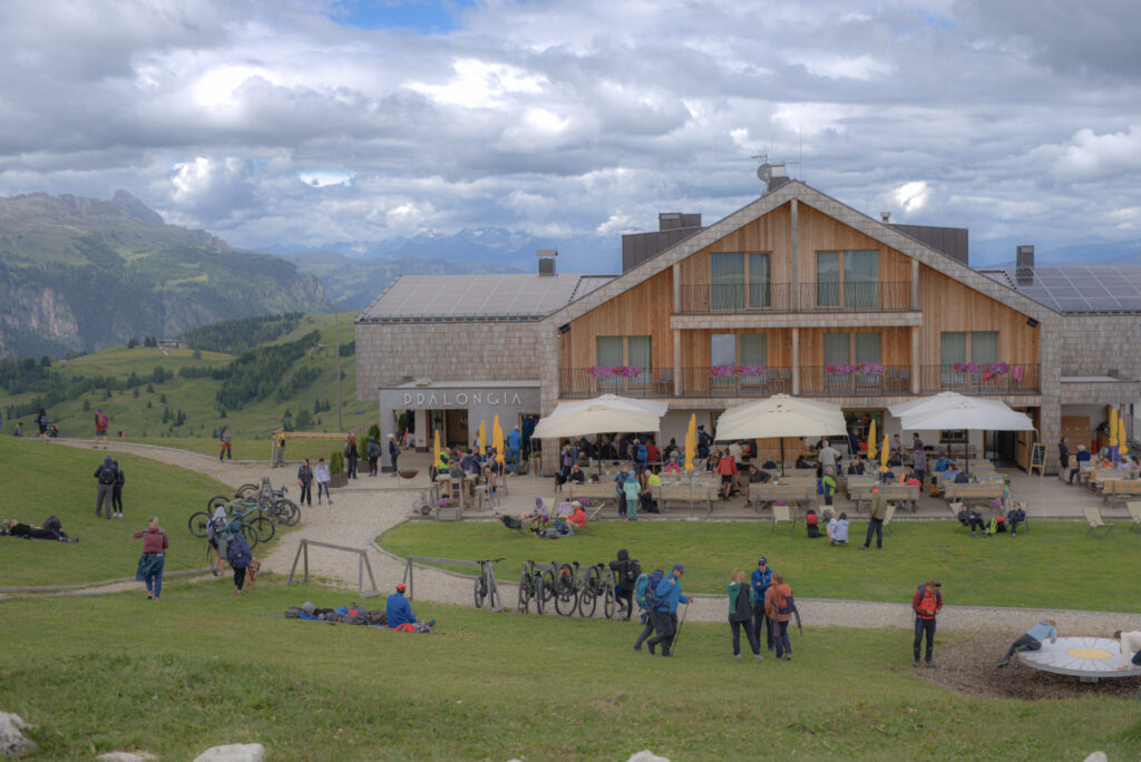 Pralongia Rifugio in Alta Badia with hikers relaxing on the grassy hillside and outdoor terrace surrounded by rolling Dolomites mountains. The wooden mountain hut labeled "PRALONGIA" sits along an easy day hike route with panoramic alpine views making it one of the best rifugios in the Dolomites for first timers.