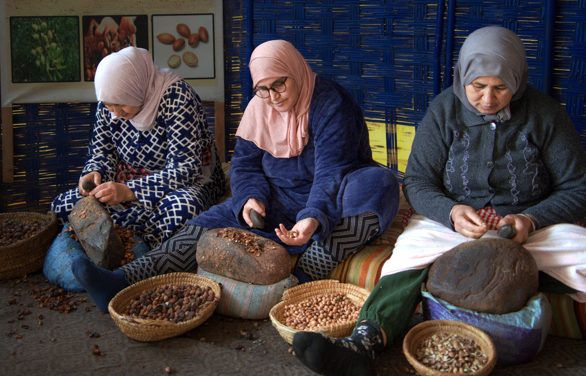 Three Berber women sit on the floor at an argan oil cooperative, hand cracking nuts between stones with woven baskets around them during a day trip from Marrakech to the Atlas Mountains.