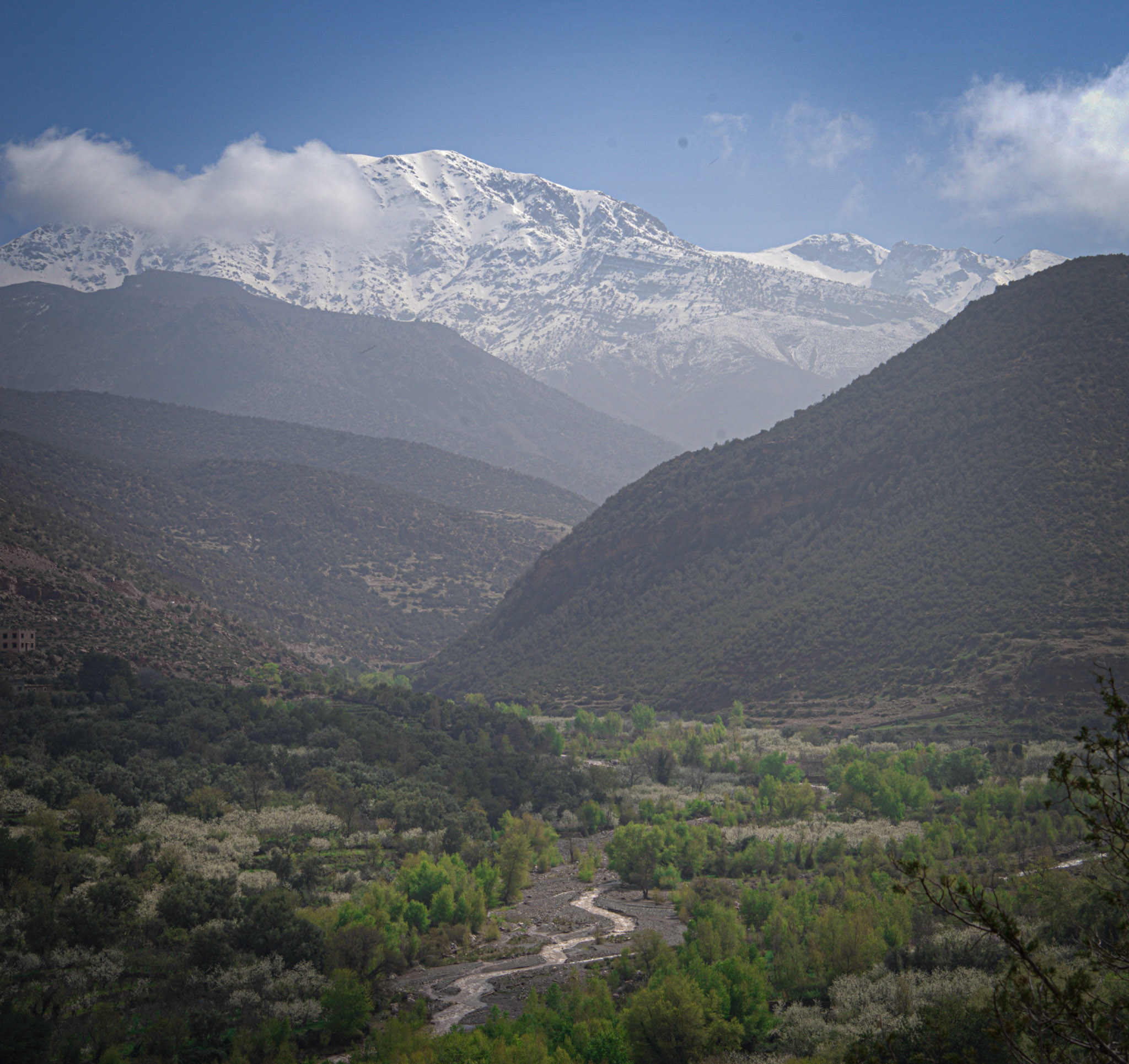 Snow capped peaks of the Atlas Mountains rise above a green valley with a winding river near Imlil, captured during a scenic hike on a Morocco itinerary.