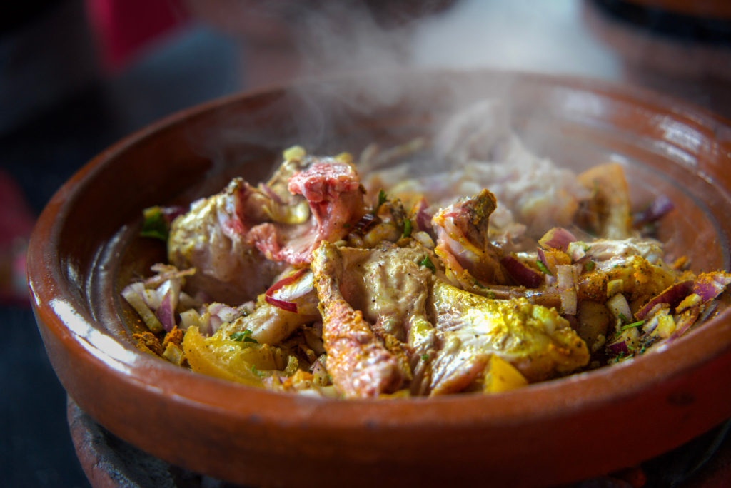 Steaming chicken tagine cooking in a clay dish with chopped onions herbs and spices during a Moroccan cooking class at the Secret Berber Garden. Pieces of chicken and vegetables simmer together as part of a traditional meal prepared during a hands on experience in Marrakech.