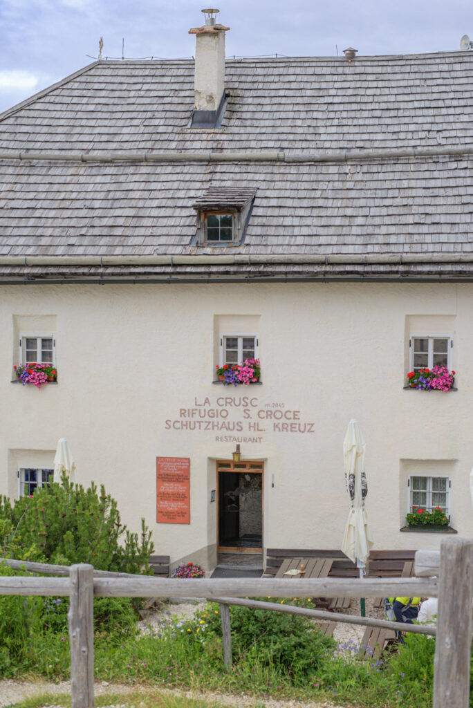 La Crusc Rifugio S Croce with white facade and flower filled window boxes set against a wooden fence and alpine greenery in the Dolomites. The building labeled "LA CRUSC RIFUGIO S CROCE SCHUTZHAUS HL KREUZ RESTAURANT" sits along an easy day hike making it one of the best rifugios in the Dolomites for first timers.