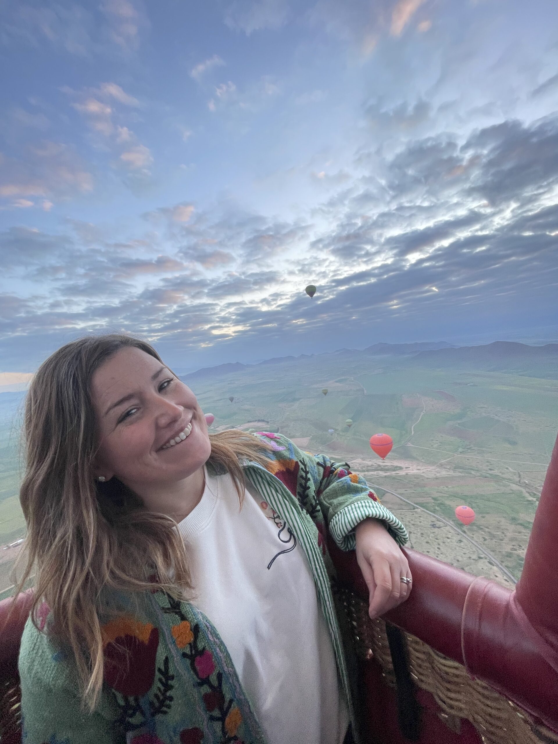 Woman smiling from a hot air balloon basket during sunrise over the desert near Marrakech with multiple balloons floating in the distance, capturing a memorable Marrakech hot air balloon ride experience.