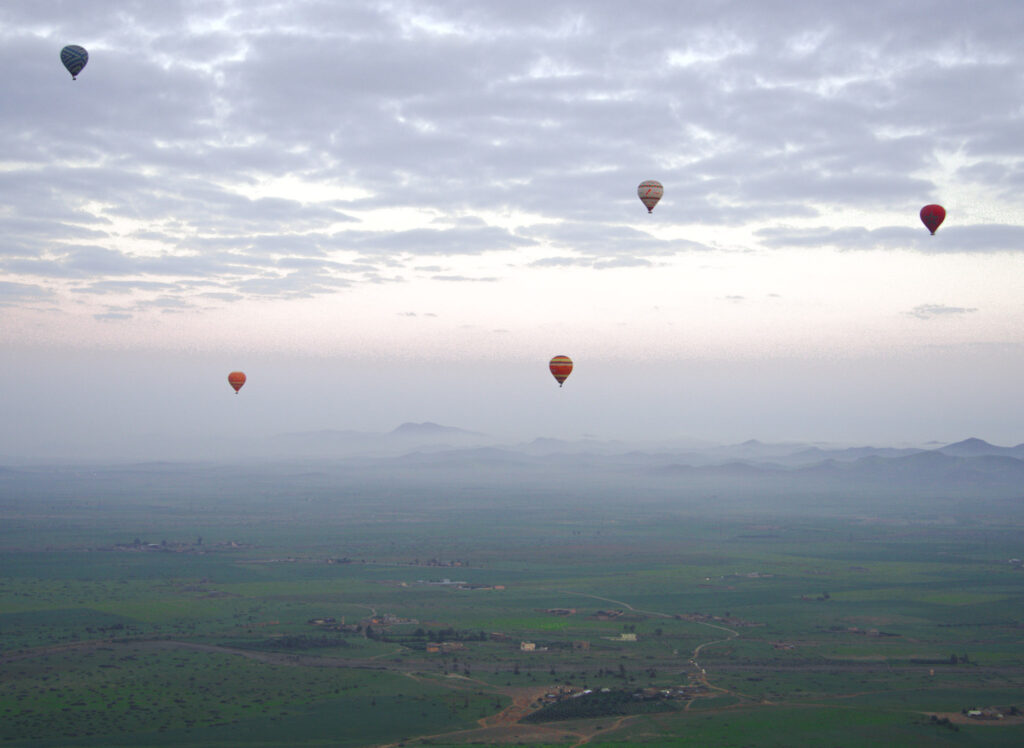 Multiple hot air balloons floating over the countryside outside Marrakech at sunrise with soft light breaking through clouds during a scenic desert ride.