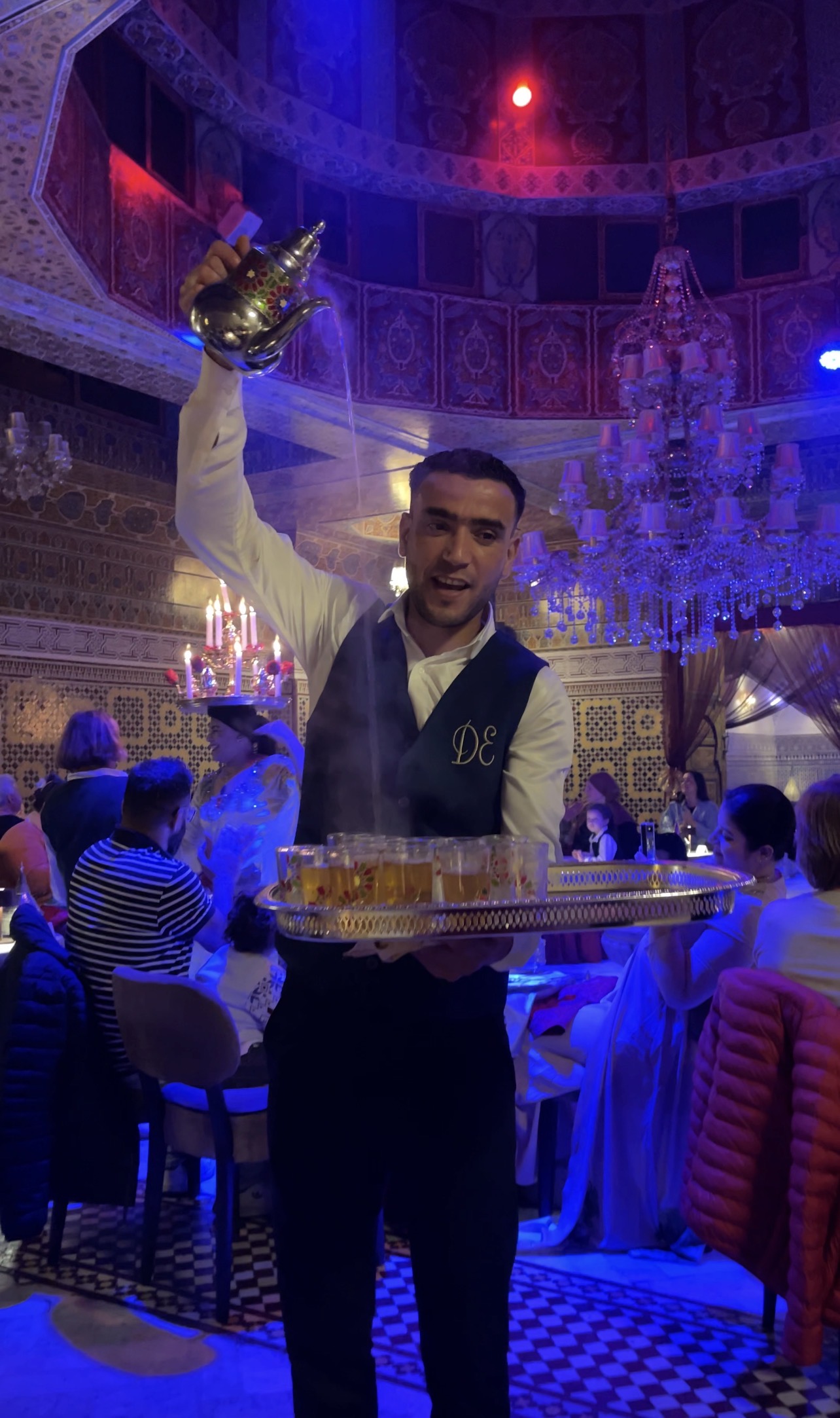 A server at Dar Essalam in Marrakech pours hot mint tea from a silver teapot into small glasses on a tray while guests sit around under colorful lighting and ornate chandeliers during a lively dinner performance.