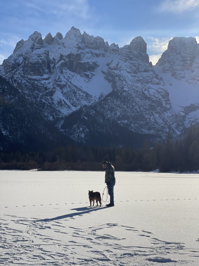 A man stands with his dog on the frozen surface of Lago di Landro in winter, surrounded by towering snow covered Dolomite mountains under a soft blue sky. Footprints trail across the ice, emphasizing the stillness and scale of the alpine landscape in December.