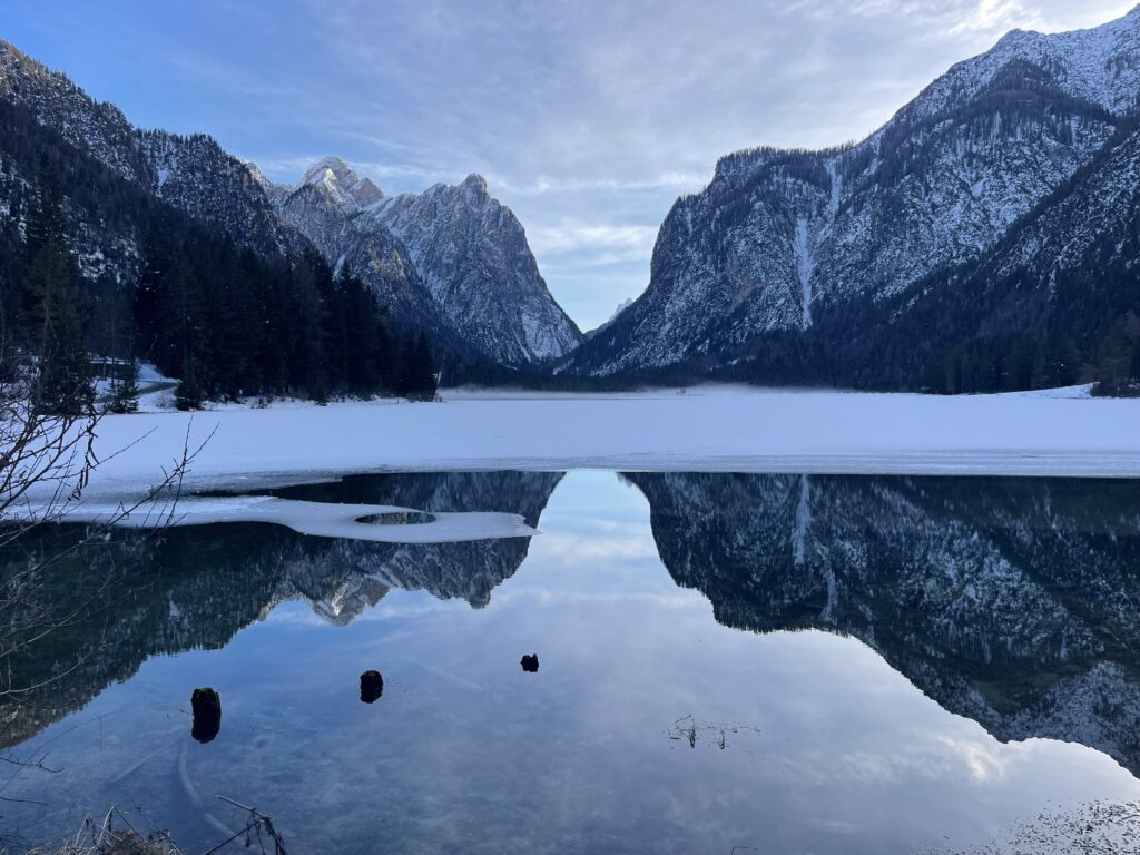he icy surface of Lago di Dobbiaco reflects dramatic Dolomite peaks on a calm December day.
Alt text: Partially frozen Lago di Dobbiaco reflects towering snow covered Dolomite mountains and pine forests in still water, with a thin layer of ice stretching across the lake. The scene is calm and symmetrical, capturing the quiet beauty of winter in the Dolomites.
