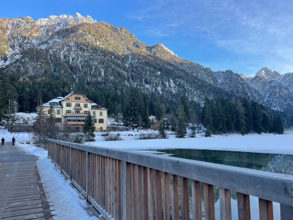 Wooden boardwalk beside the frozen edge of Lago di Dobbiaco in winter with a pale yellow lodge nestled among pine trees and snow covered mountains in the background under a clear blue sky. A few people walk along the path, adding scale to the quiet alpine setting.