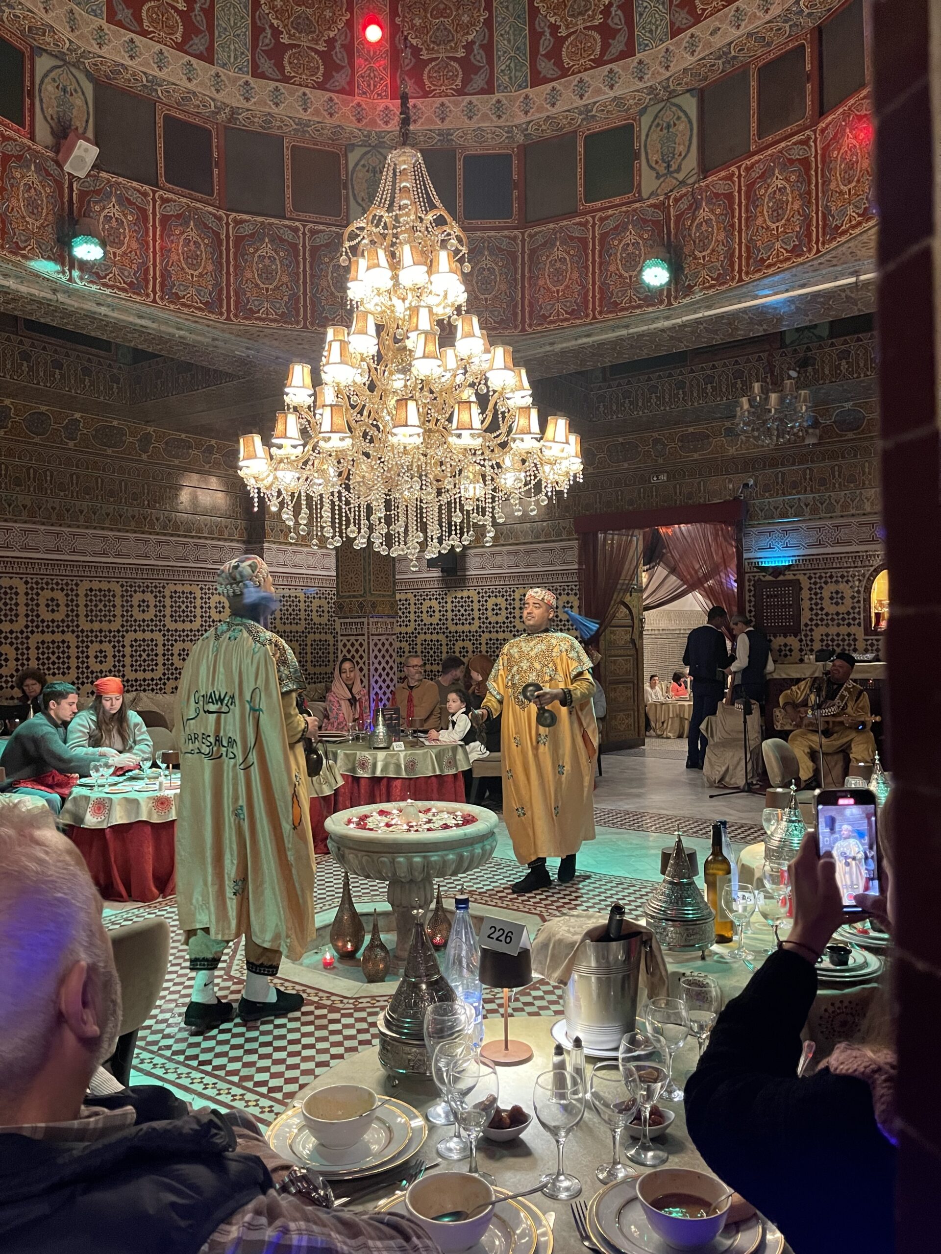 Musicians in traditional Moroccan clothing perform in the center of Dar Essalam in Marrakech beneath a large chandelier while diners sit around tables watching the live entertainment during dinner.