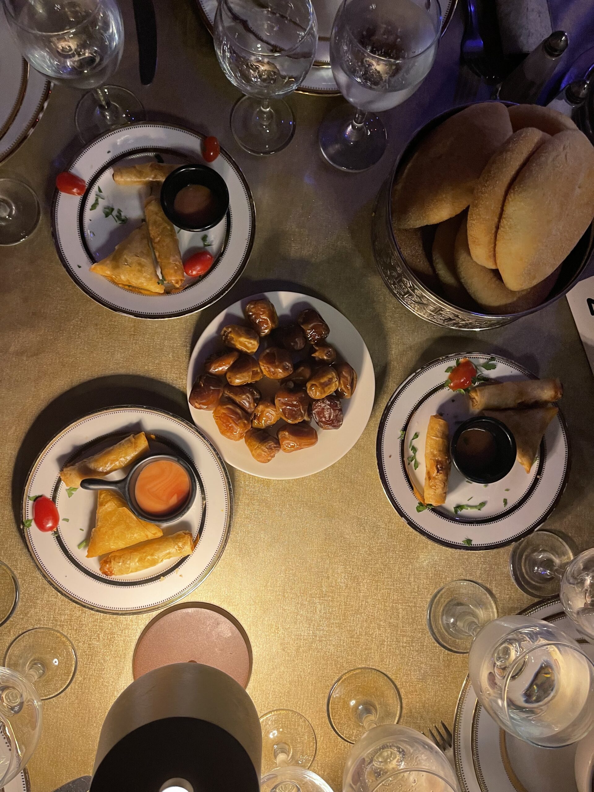 A table at Dar Essalam in Marrakech set with traditional starters including crispy pastries with dipping sauce, a bowl of dates, round bread, and multiple glasses arranged for a shared dinner experience.