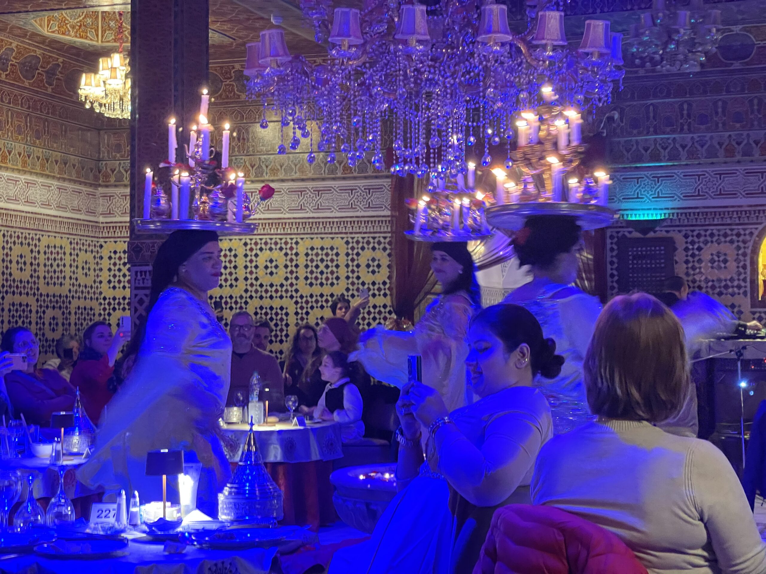 omen in flowing dresses balance trays of lit candles on their heads while dancing through the dining room at Dar Essalam in Marrakech as guests watch and take photos during an evening dinner show.