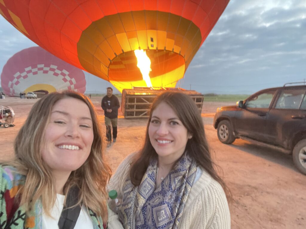 Two women smiling in front of a glowing hot air balloon being inflated on the ground in Marrakech at sunrise, showing the start of a hot air balloon Marrakech adventure and one of the most unique things to do in Marrakech.