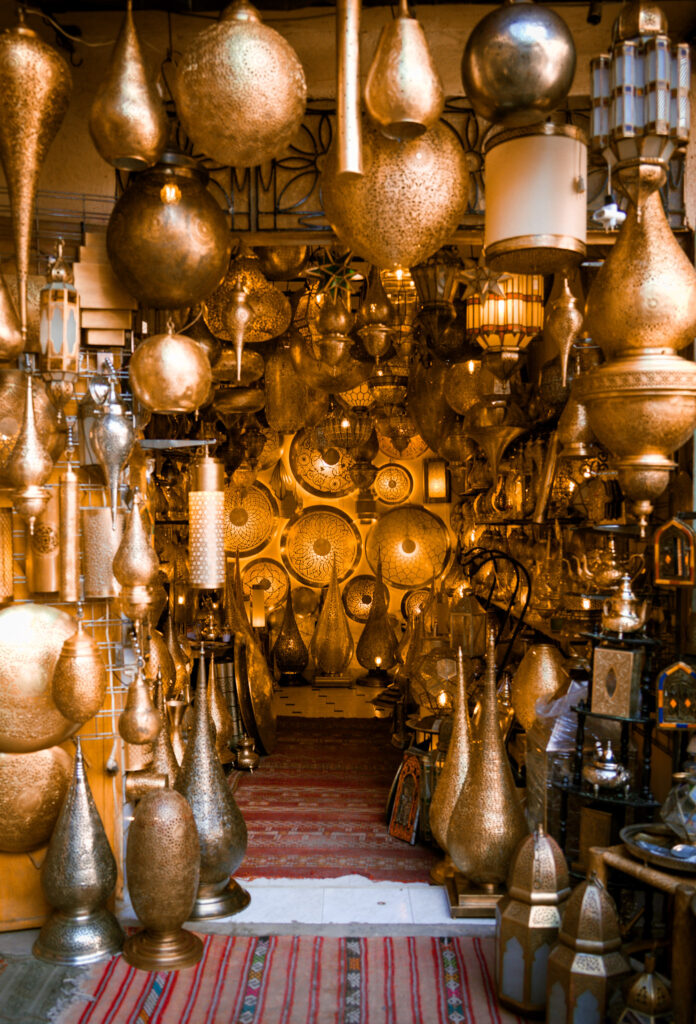Narrow souk shop in Marrakech filled with hanging brass lanterns and intricately patterned metal decor glowing with warm golden light during a medina walking tour.