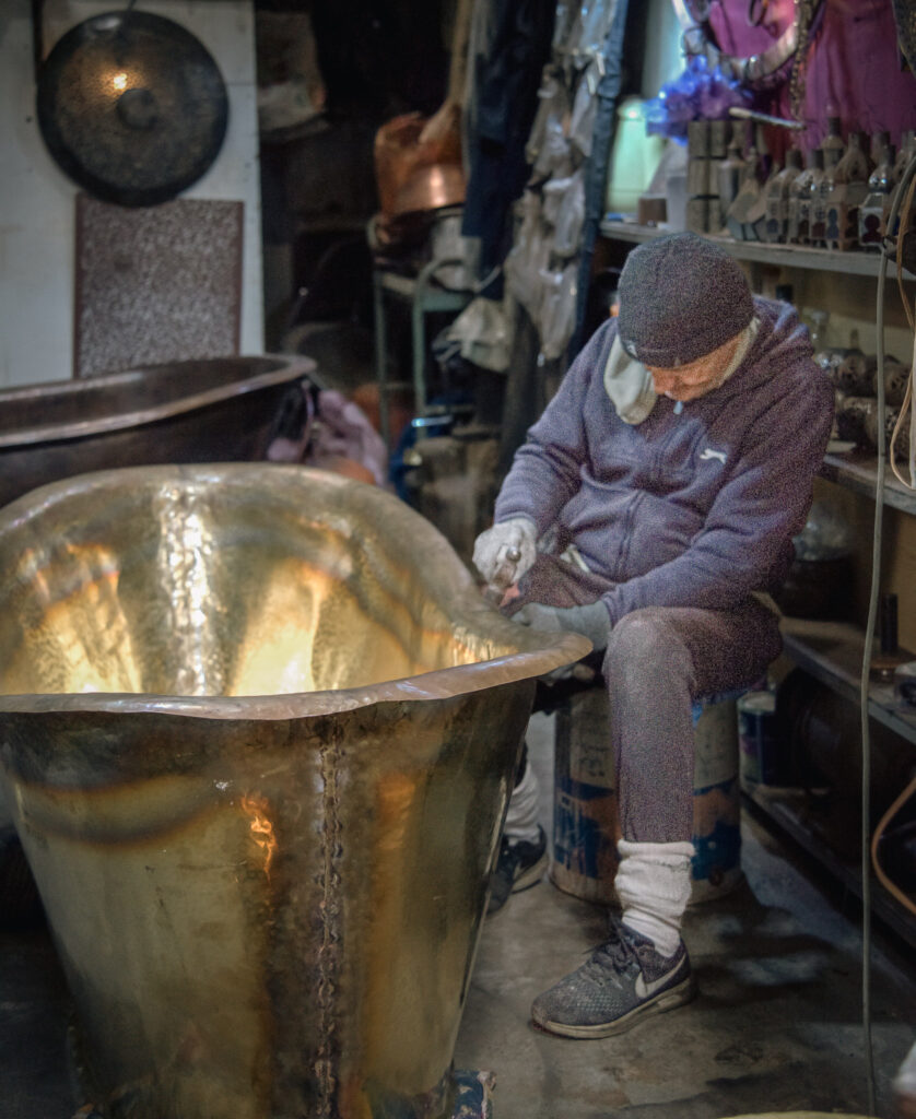 Artisan in a Marrakech workshop carefully shaping a large hammered metal tub by hand, showing traditional craftsmanship found while exploring the souks.