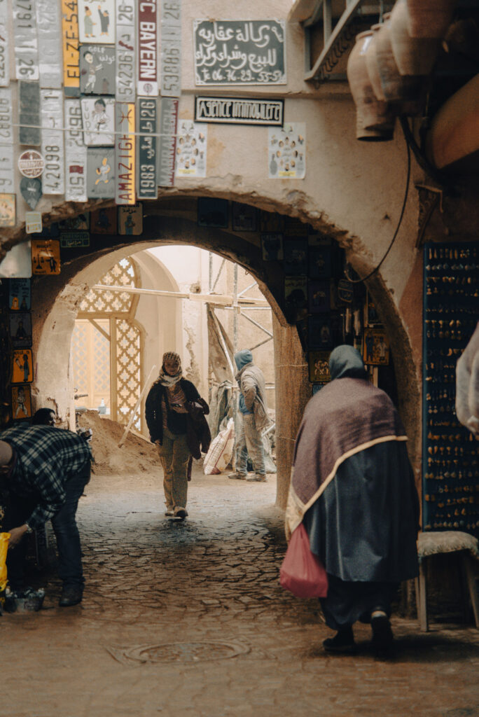Arched passageway in the Marrakech medina with people walking through a busy souk street lined with signs, crafts, and textured walls.