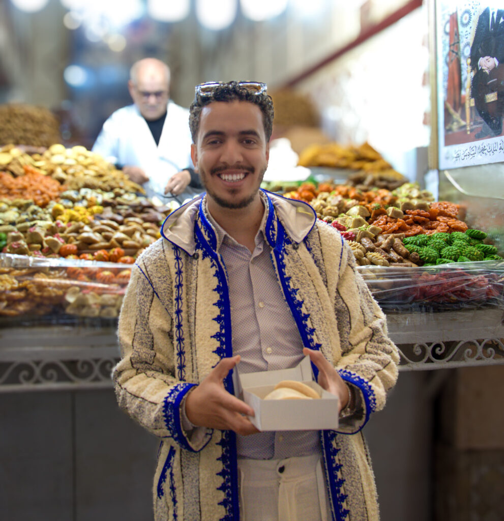Smiling vendor in a Marrakech souk holding a box of sweets with colorful market stalls full of pastries and dried goods behind him.
