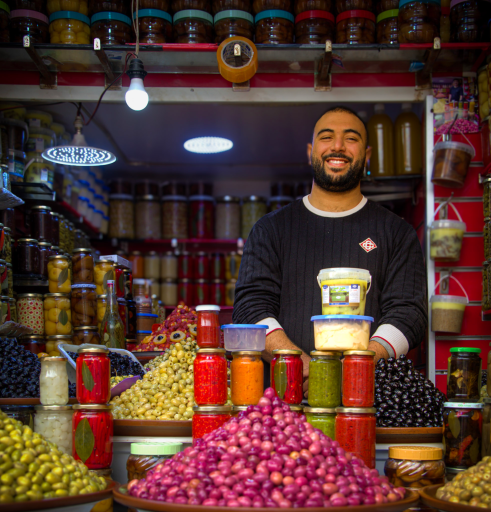 Market stall in the Marrakech souks displaying piles of olives and jars of preserved vegetables while a vendor stands behind the counter smiling.