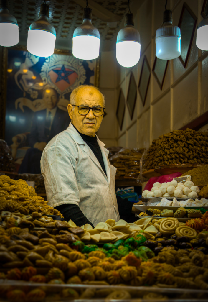 Older shopkeeper standing behind a display of traditional Moroccan pastries and sweets under hanging lights inside a Marrakech souk.