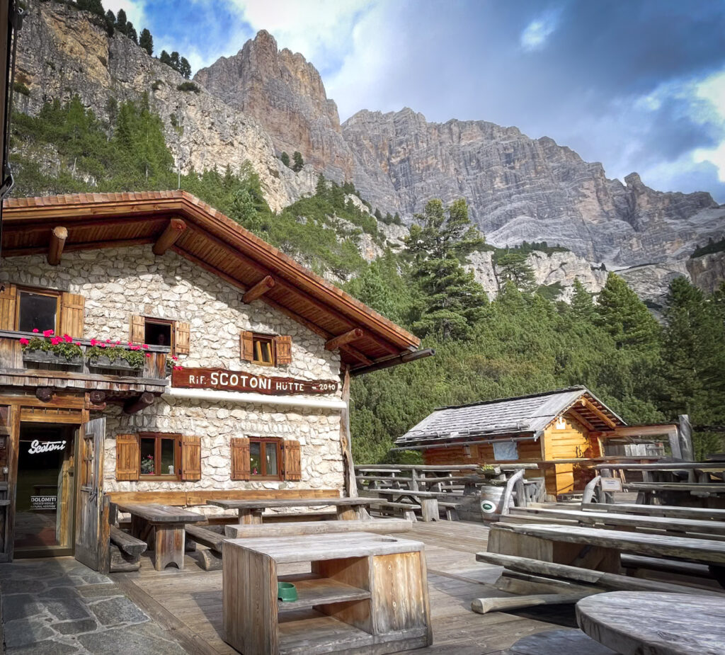 Rifugio Scotoni mountain hut with wooden terrace seating beneath towering Dolomites cliffs along the Alta Via 1 hiking route. The rustic stone building labeled "Rif. SCOTONI HÜTTE" sits in a forested alpine setting making it one of the best rifugios in the Dolomites on an easy day hike for first timers.