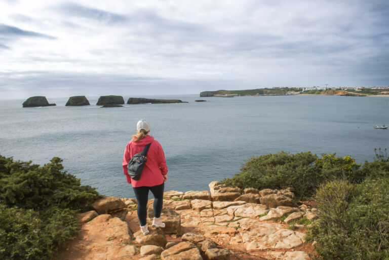 Alt text: Woman in a red jacket stands on a rocky cliff overlooking the ocean and small offshore rock formations near Sagres, capturing the scenic coastal views highlighted in this hotel in the Algarve review.