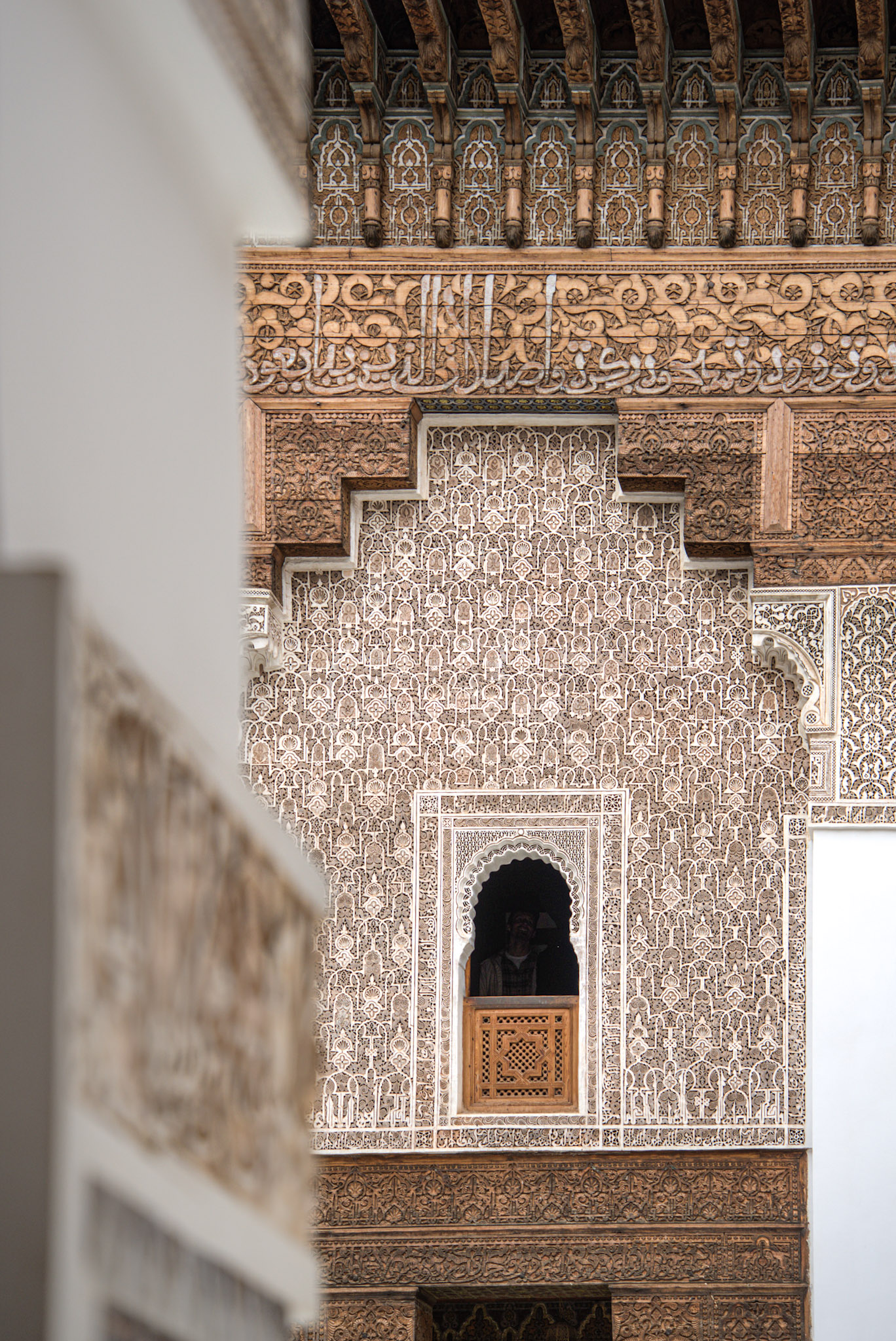 Ornate courtyard wall of Ben Youssef Madrasa in Marrakesh framed by a blurred arch in the foreground. A person stands in a carved window niche surrounded by dense geometric patterns, showcasing the historic architecture featured in things to do in Marrakesh.