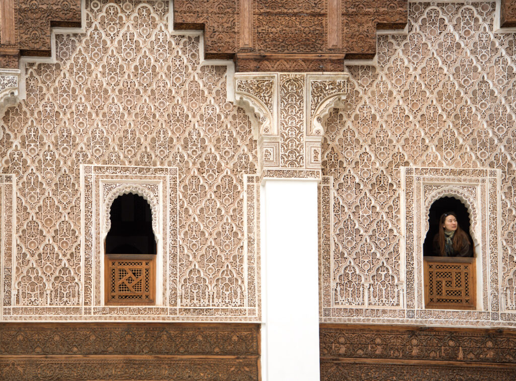 Intricately carved walls inside Ben Youssef Madrasa in Marrakesh with repeating geometric and floral patterns. A woman stands in a small arched window looking out, highlighting the detailed craftsmanship visitors can admire when exploring things to do in Marrakesh.
