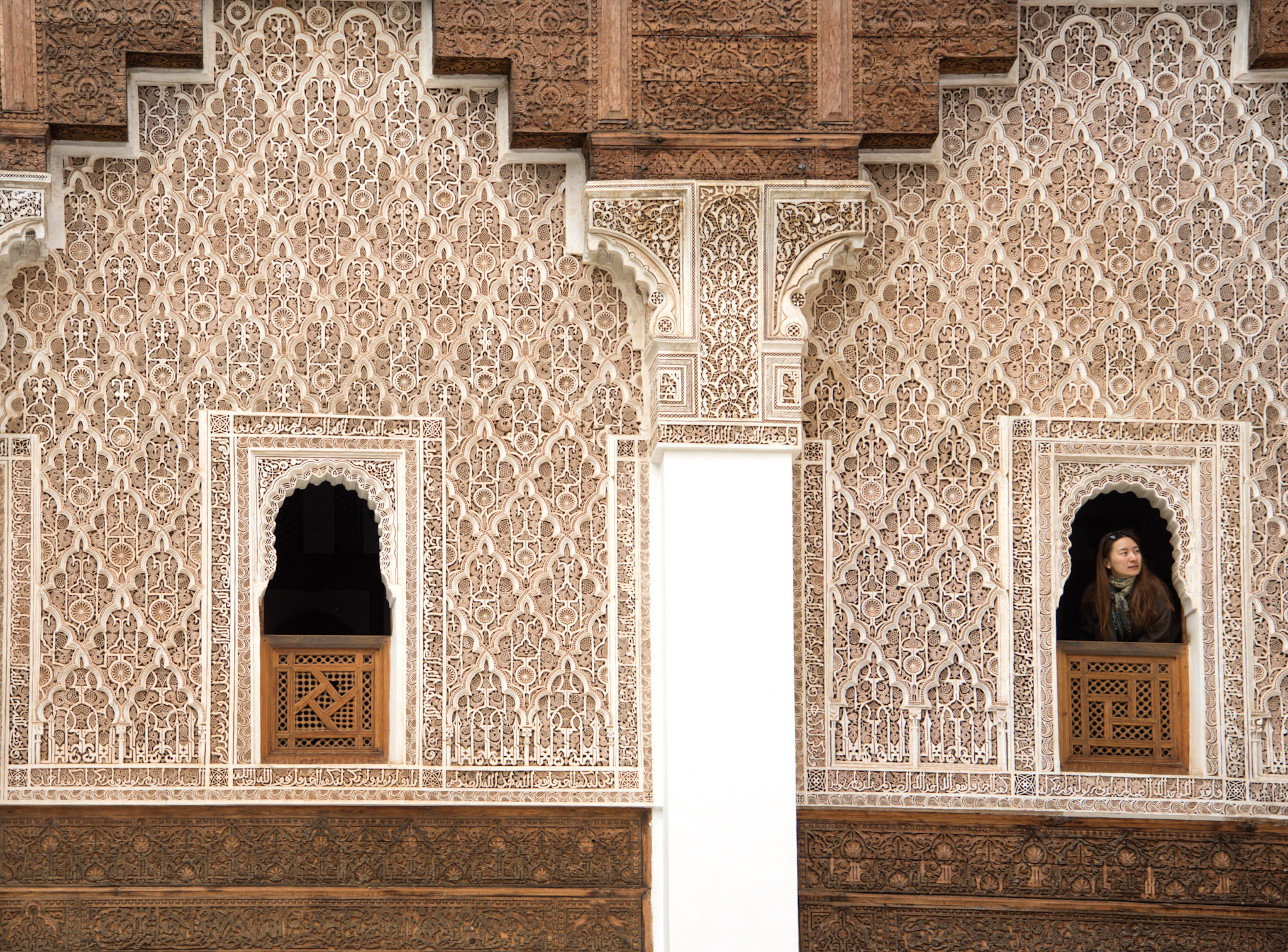 Intricately carved walls inside Ben Youssef Madrasa in Marrakesh with repeating geometric and floral patterns. A woman stands in a small arched window looking out, highlighting the detailed craftsmanship visitors can admire when exploring things to do in Marrakesh.