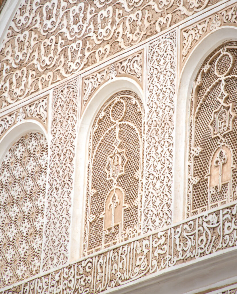 Close up of carved stucco and wood detailing at Ben Youssef Madrasa in Marrakesh with Arabic calligraphy running across the wall. Repeating arches and intricate patterns demonstrate the artistry that makes this site a highlight among things to do in Marrakesh.