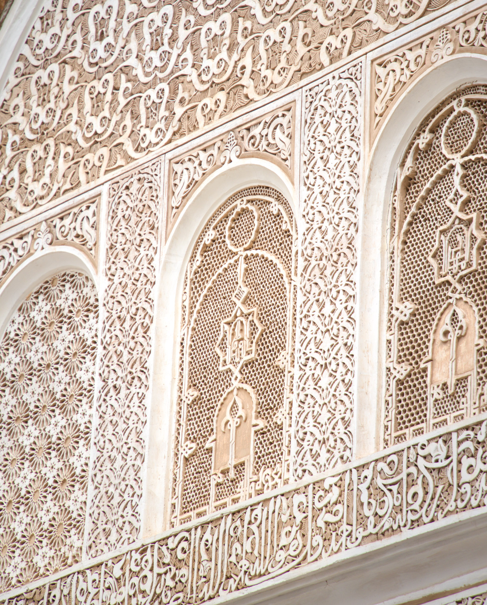 Close up of carved stucco and wood detailing at Ben Youssef Madrasa in Marrakesh with Arabic calligraphy running across the wall. Repeating arches and intricate patterns demonstrate the artistry that makes this site a highlight among things to do in Marrakesh.