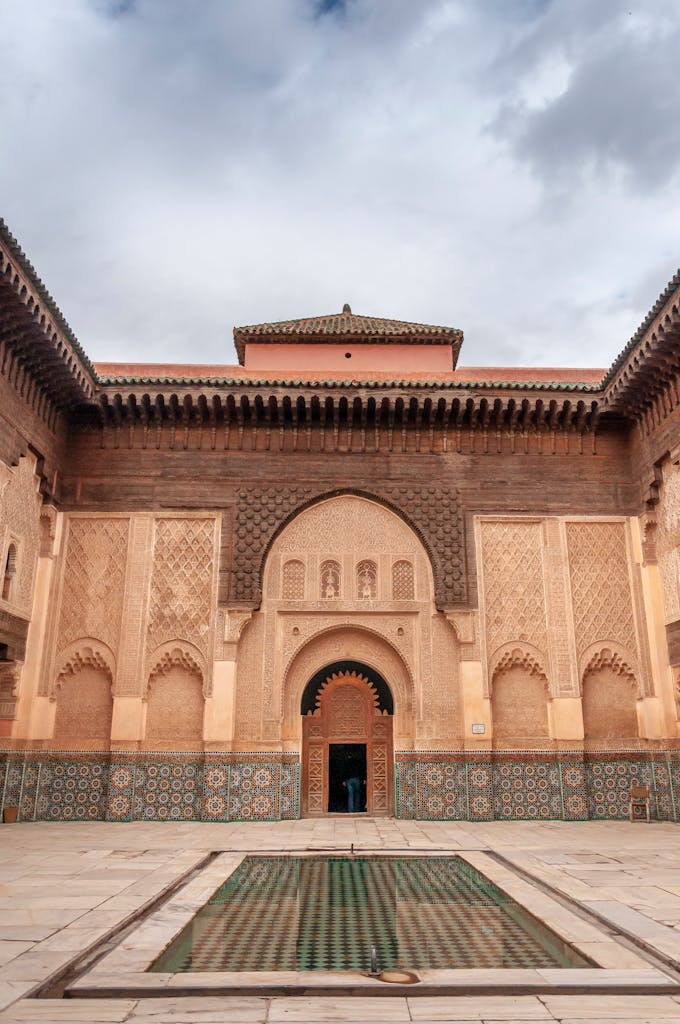 Water basin in courtyard of ancient Ben Youssef Madrasa with ornamental walls decorated with arabesque and zellij tilework against overcast sky in Marrakesh