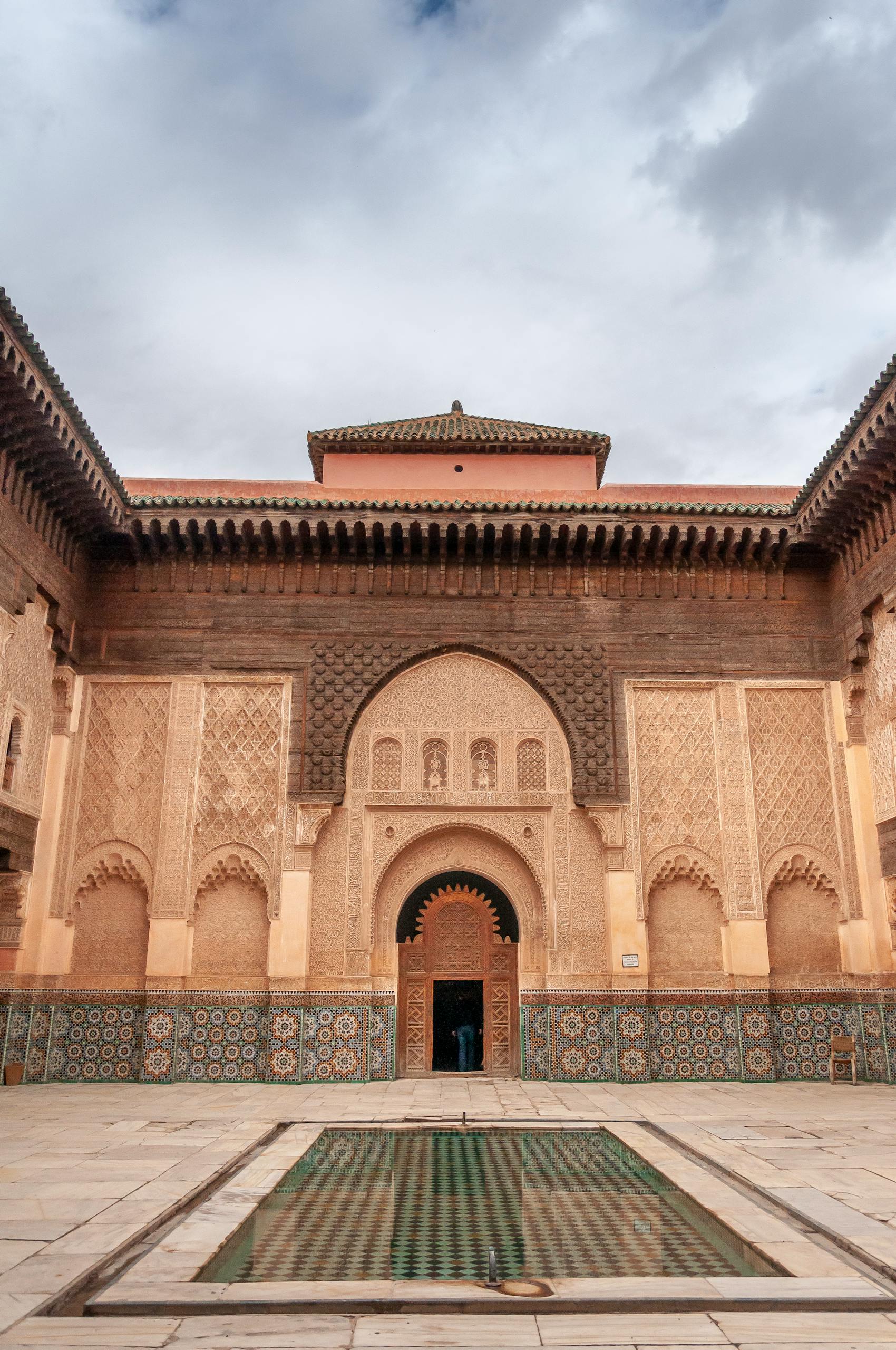 Water basin in courtyard of ancient Ben Youssef Madrasa with ornamental walls decorated with arabesque and zellij tilework against overcast sky in Marrakesh