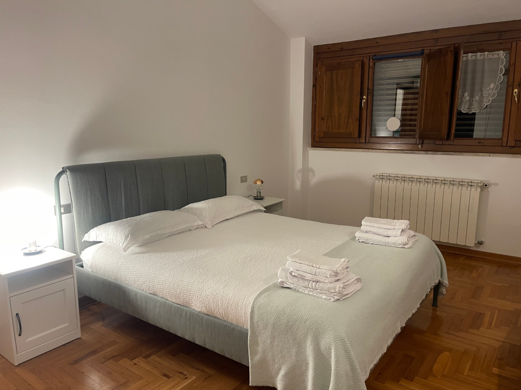 Minimalist bedroom in a Siena apartment with a large bed white linens folded towels and a window with wooden shutters. This comfortable space offers a relaxing stay during a 2 days in Siena itinerary.