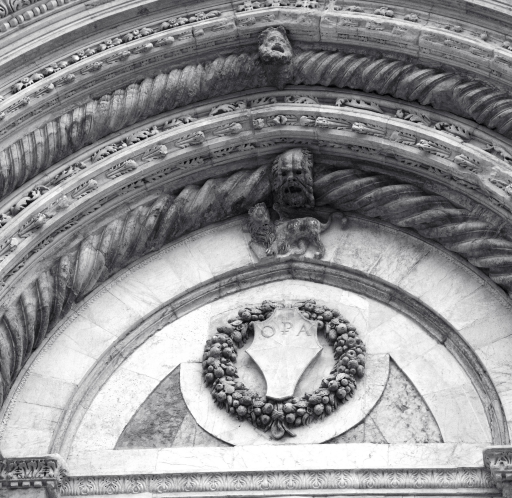 Black and white image of marble relief portraits of bearded men and lions set in diamond panels on the Siena Duomo facade.