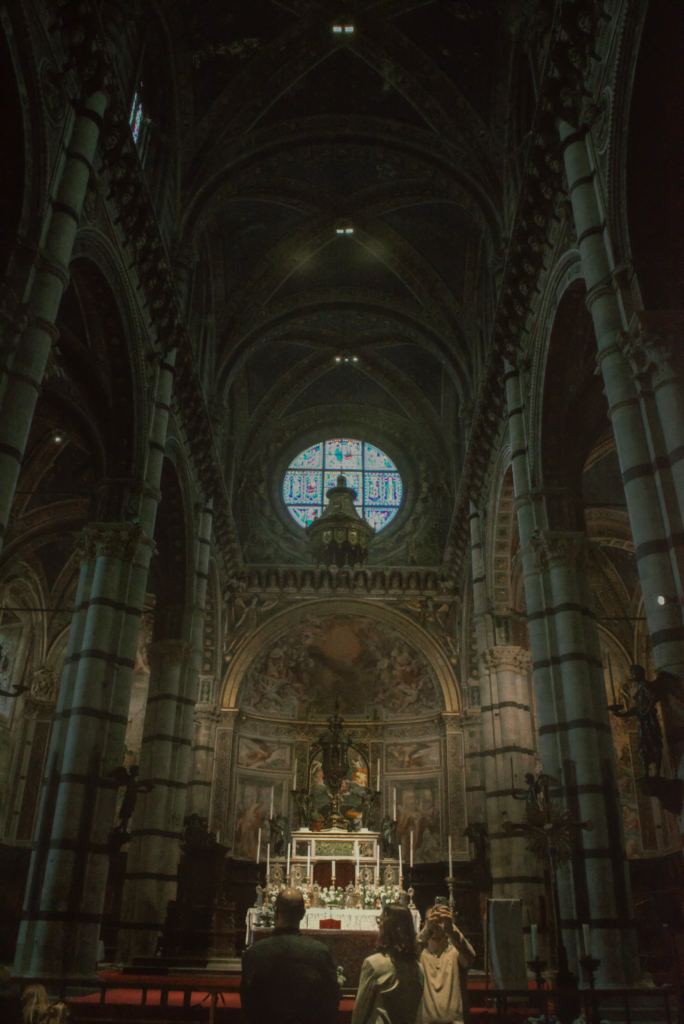 Wide view of the Siena Duomo interior showing the altar beneath a large circular stained glass window. Visitors stand quietly in the dimly lit space surrounded by towering columns.