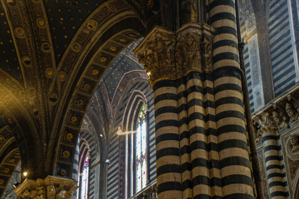 Alt text: Interior of the Siena Duomo with tall striped columns and arched ceilings covered in gold stars. Light filters through stained glass windows creating a dramatic atmosphere inside the cathedral.