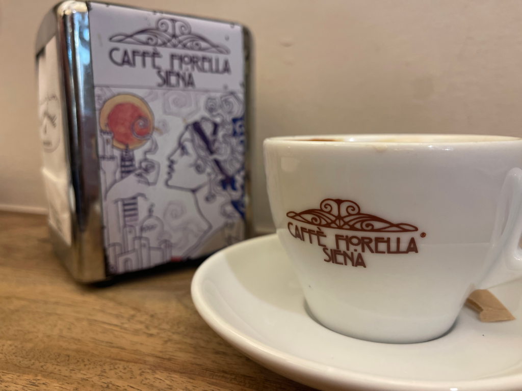 Espresso cup on a saucer with the text "CAFFÈ FIORELLA SIENA" beside a matching napkin holder on a wooden table. This cozy cafe moment captures a typical break during a 2 days in Siena itinerary.