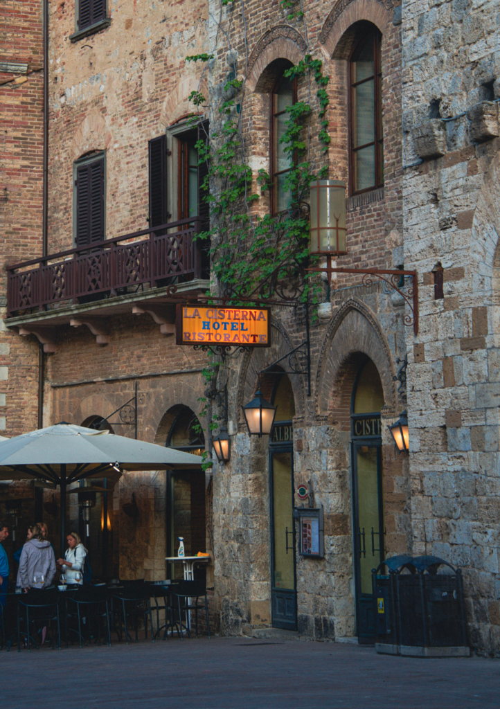 Stone arches and brick walls frame a restaurant terrace in a medieval town square. A yellow sign reads “LA CISTERNA HOTEL RISTORANTE.
